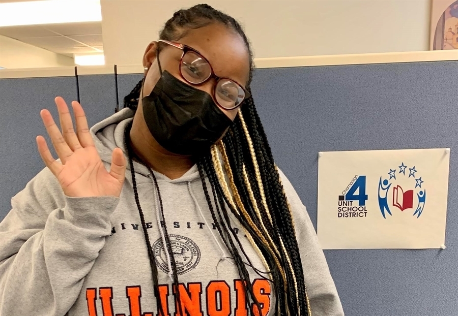 An masked tutor with glasses and long black braids  in a University of Illinois hoodie smiles and waves at the camera